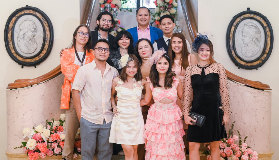 Group of people posing in formal attire with a floral arrangement and classical portraits in the background.