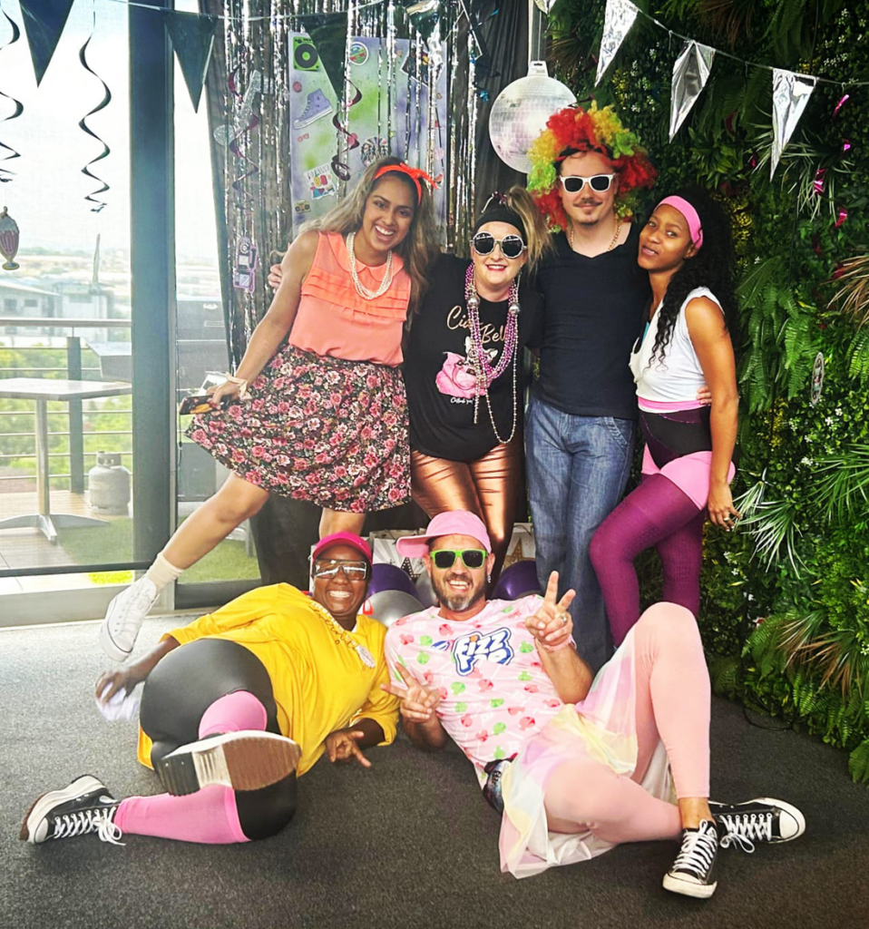 Group of people in colorful outfits posing playfully at a party with decorations.