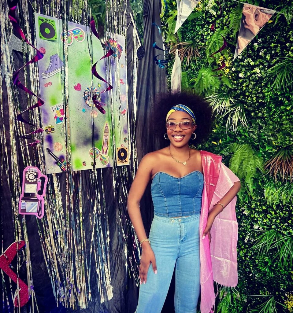 Person in denim outfit standing in front of a mixed backdrop with a retro and greenery theme.