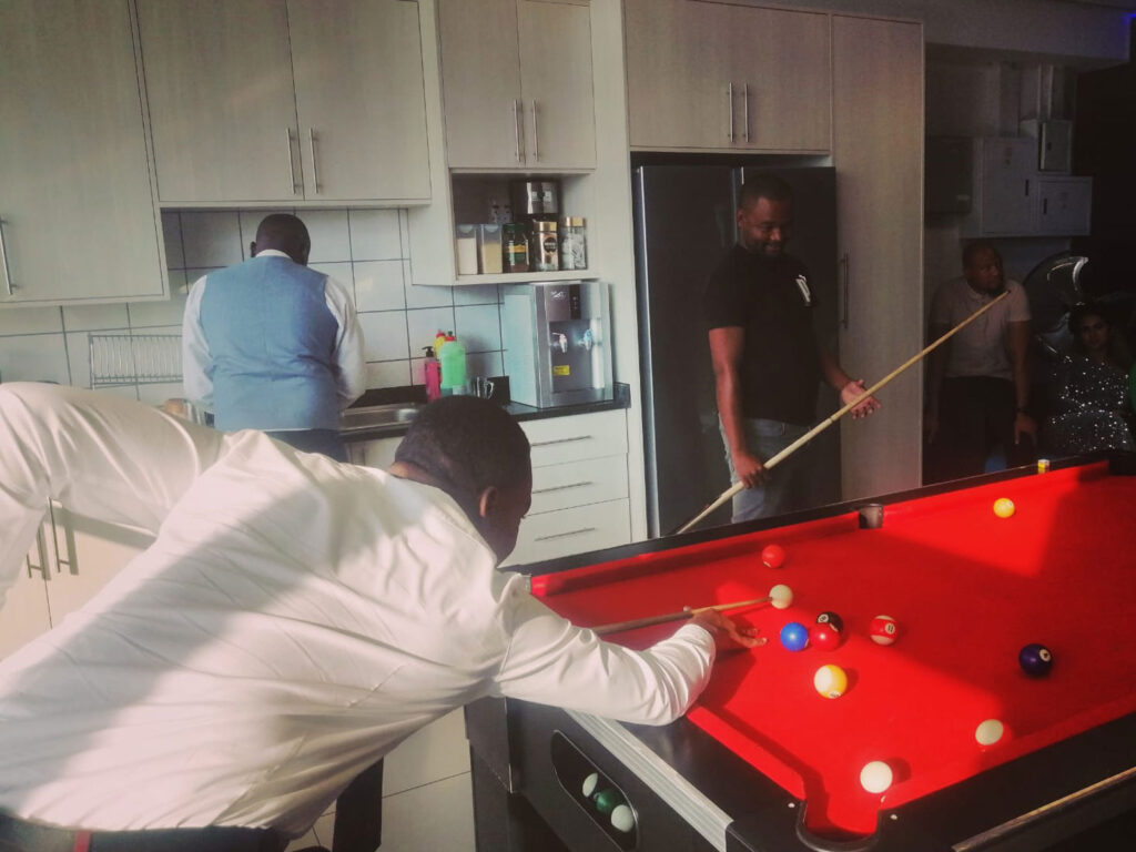 Man in white shirt playing pool with red table, others in background.