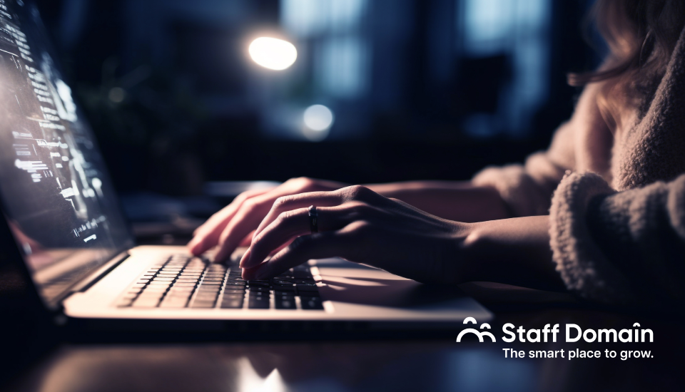 Person typing on a laptop in a dimly lit room with code on screen and "Staff Domain" logo in corner.