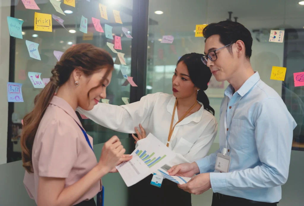 Three professionals discussing charts, with sticky notes on glass wall behind them.