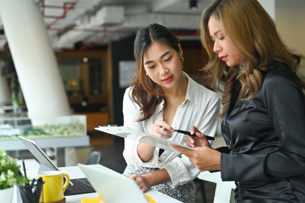 Two women discussing over documents next to a laptop in an office setting.