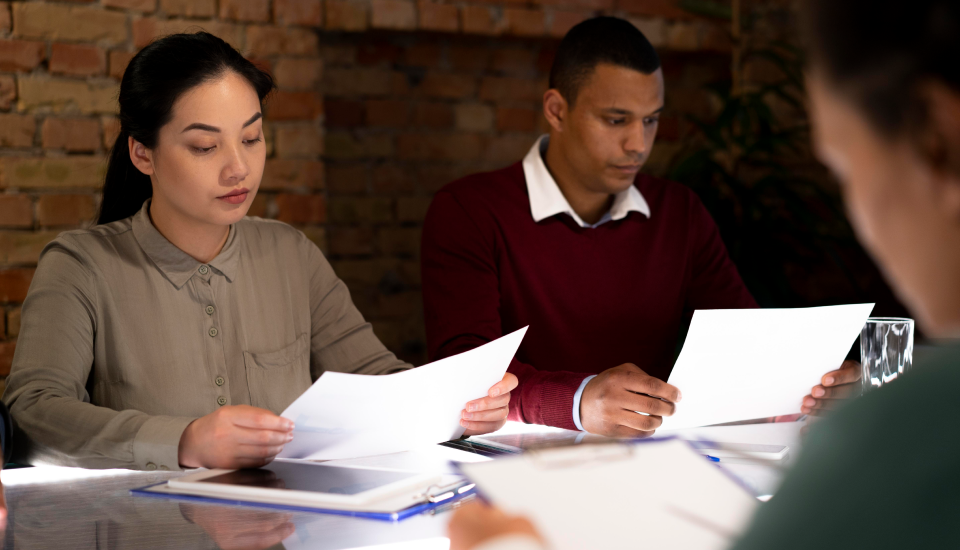 Two individuals sitting at a table reviewing documents in a meeting room with a brick wall in the background.