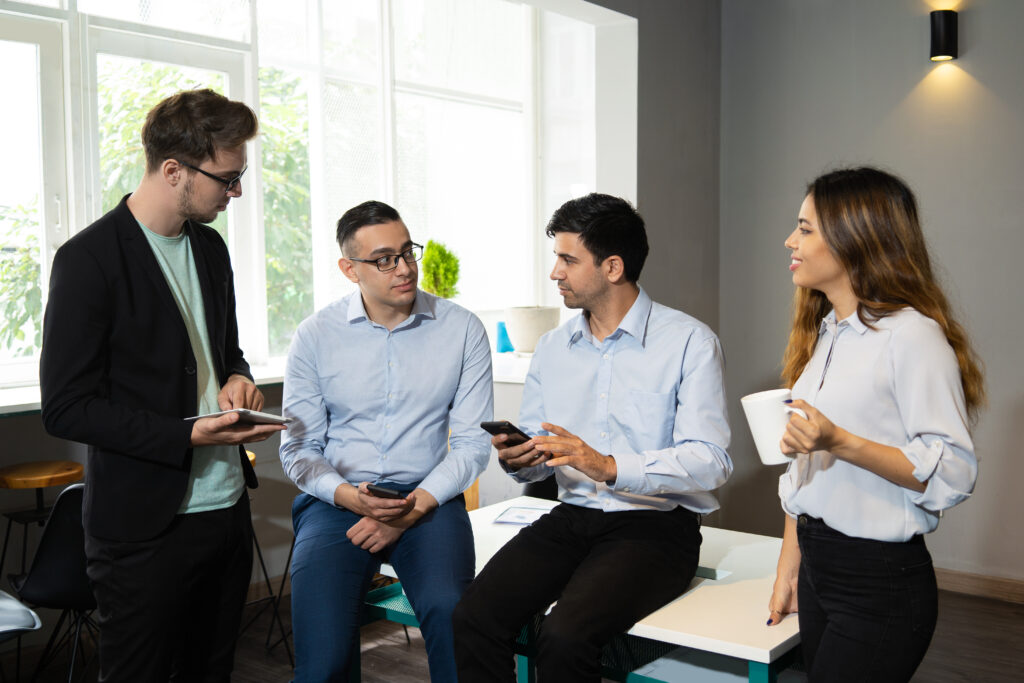 Professional team consulting on internet while discussing ongoing project. Four employees talking in meeting workspace, using gadgets and drinking coffee. Interaction concept